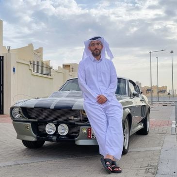 Man in traditional attire leaning on a classic car outdoors.