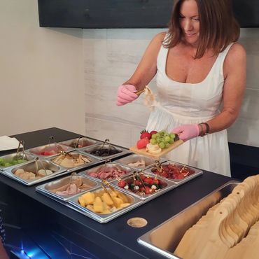 Woman in white dress preparing a charcuterie board with gloves at a Luxe Provision & Co. counter.