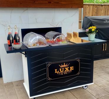 Outdoor black serving cart with food covers and drinks, branded Luxe Provisions & Co.