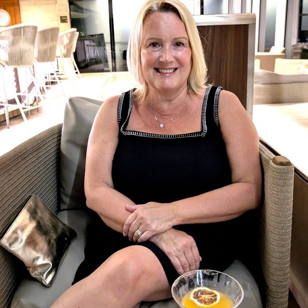 Smiling woman in black dress sitting on a chair with a drink.