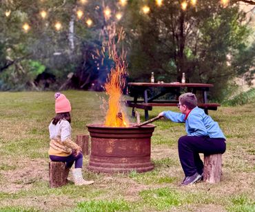 Two children sit on logs by a fire pit outdoors at dusk.