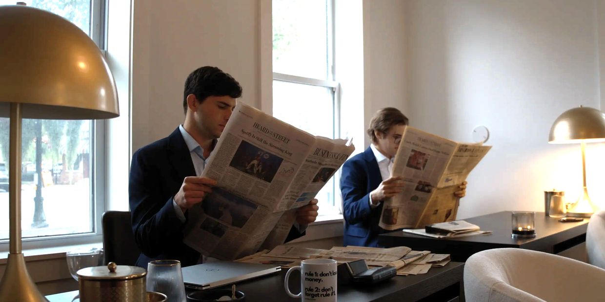 Two men in suits reading newspapers at an office desk.
