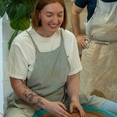 A smiling woman shaping clay on a pottery wheel wearing a gray apron.