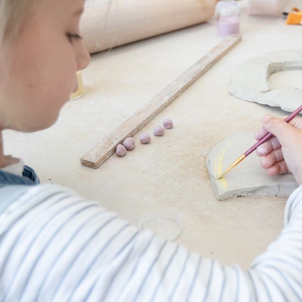 Child painting a clay piece with yellow paint on a worktable.