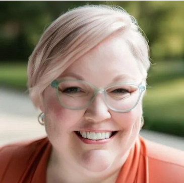 Smiling woman with short blond hair, light blue glasses, and orange blazer.