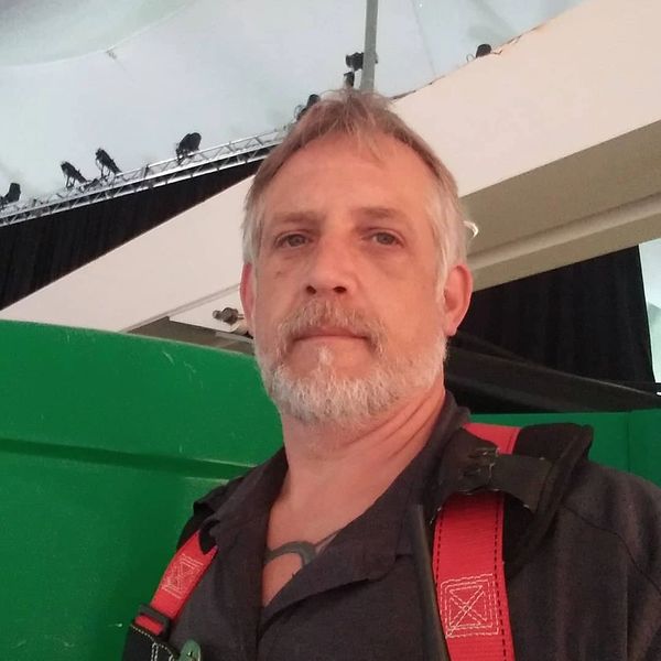 Middle-aged man with gray hair and beard wearing a dark shirt and red suspenders indoors.