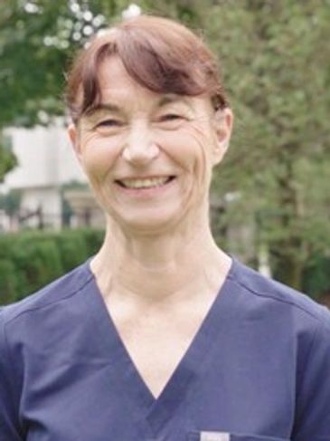 Smiling woman in blue scrubs outdoors with greenery background.