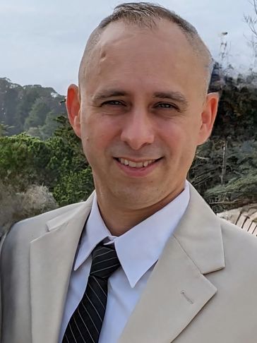 Man in beige suit and black striped tie smiling outdoors.