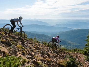 Two mountain bikers ride down a steep, rocky trail with scenic mountain views.
