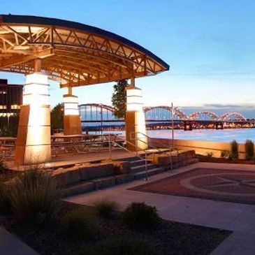 Illuminated riverside pavilion with a bridge in the background at dusk.