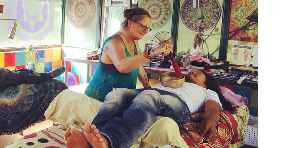 A woman performs a sound healing session using a singing bowl on a man lying on a bed.