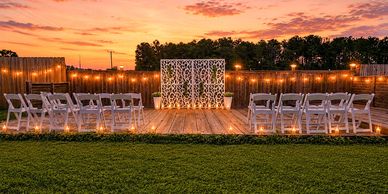 Outdoor wedding setup with white chairs and decorative backdrop at sunset.