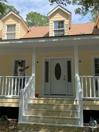Person standing on the porch of a yellow house with white railings.