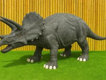 A realistic triceratops dinosaur model on green grass with a bamboo fence background.