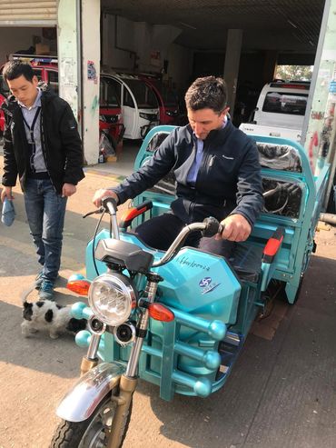 Man testing a turquoise three-wheeled vehicle while another man and a dog stand nearby.