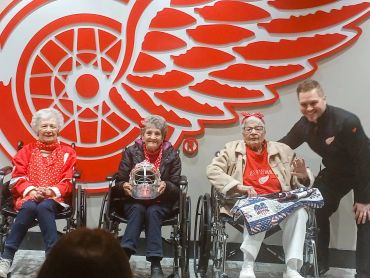 Three elderly women in wheelchairs with a man posing in front of a large Detroit Red Wings logo.