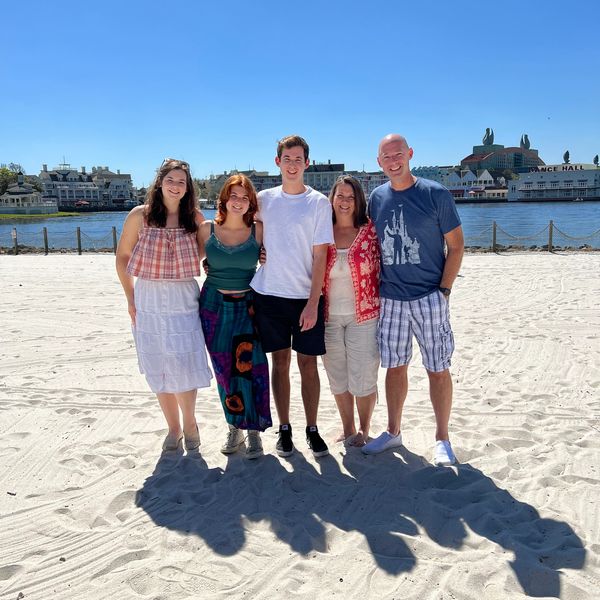 A happy family of five posing on a sunny beach with water and buildings in the background.