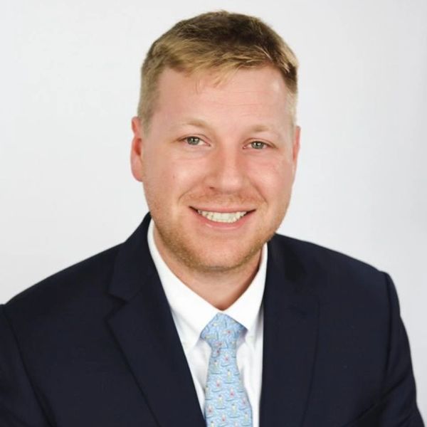 Smiling man in a navy suit and light blue patterned tie against a plain background.