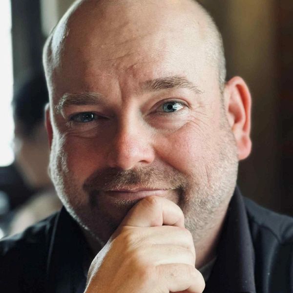 Close-up portrait of a bald man with blue eyes, smiling thoughtfully.