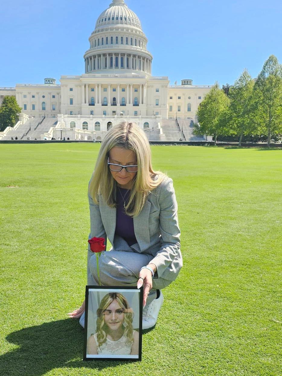 Woman kneels on grass holding a framed photo and a red rose in front of the U.S. Capitol.