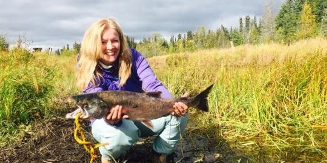 A woman proudly holds a large fish in a grassy outdoor setting.