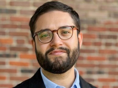 A man with glasses and a beard smiling in front of a brick wall.
