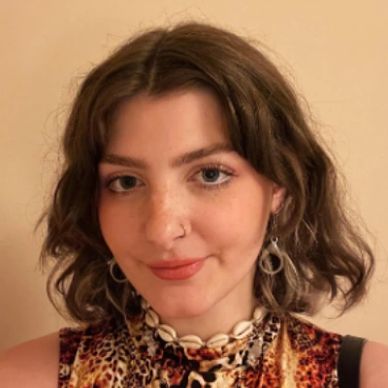 Young woman with wavy brown hair wearing a patterned top and shell necklace.