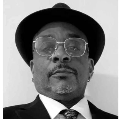 Man in glasses, hat, and formal suit looking at the camera.