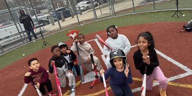 Seven kids posing with baseball bats on a field, smiling and making peace signs.