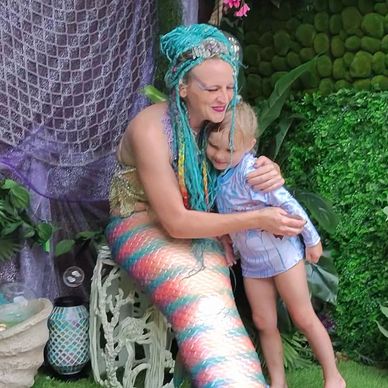 A woman dressed as a mermaid hugs a young child in a garden setting.
