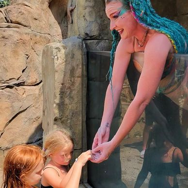 A performer with blue hair interacts with two children at an aquarium or zoo.