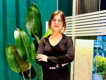 A woman in black dress standing confidently indoors near green plants.