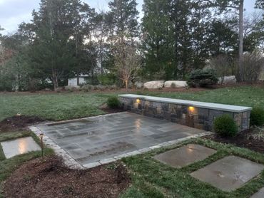 A stone patio with a built-in stone bench and soft lighting in a backyard.