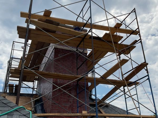 Scaffolding around a brick chimney on a roof under cloudy sky.
