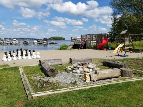 Lakeside playground with giant chess, fire pit, and slide under blue sky.