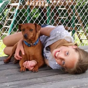 A girl lying on a wooden deck hugging a small brown dog.