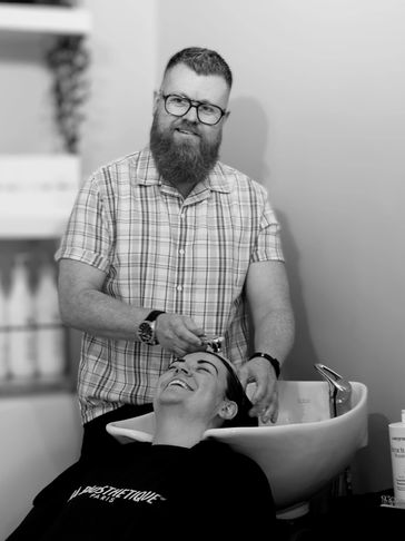 Barber washing a client's hair in a salon, both smiling warmly.