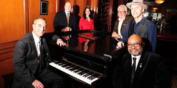 The Mike Petrone Band posing in front of a shiny piano in an oak room.