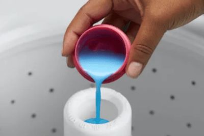 Hand pouring blue liquid detergent into a washing machine dispenser.