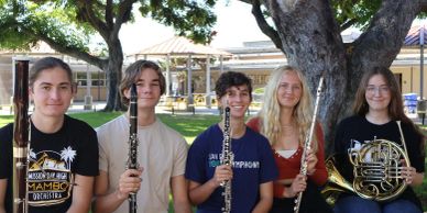 Four young musicians sitting outdoors holding woodwind and brass instruments.