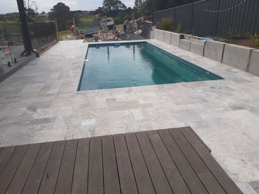 Rectangular backyard pool with stone tiles and wooden deck under construction.