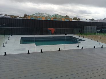 Modern backyard pool with glass fencing and deck under cloudy sky.