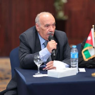 An elderly man in a suit speaks into a microphone at a conference table.