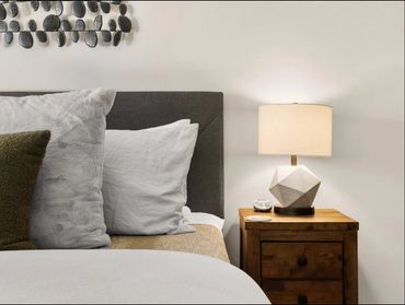 Cozy bed with pillows and a wooden nightstand holding a modern geometric lamp.