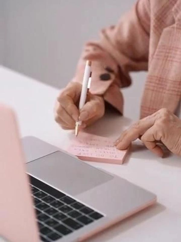 Person writing notes on a pink sticky pad next to a laptop.