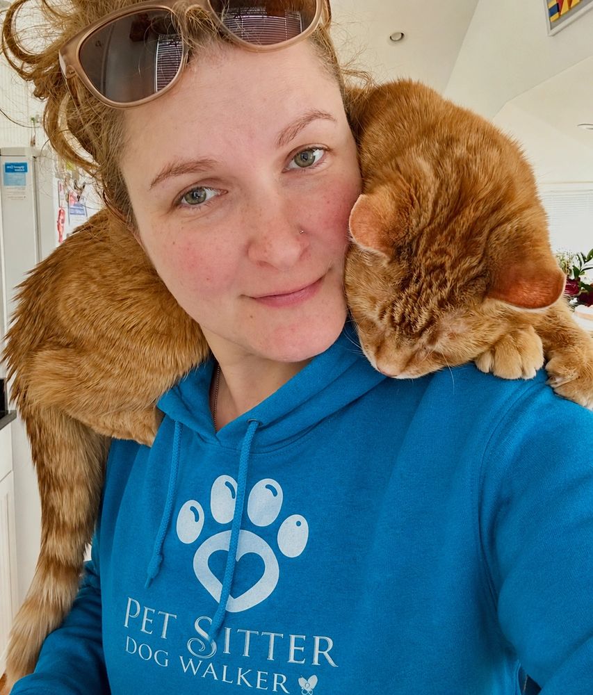 Woman with curly hair and sunglasses on her head, smiling with an orange cat on her shoulder.