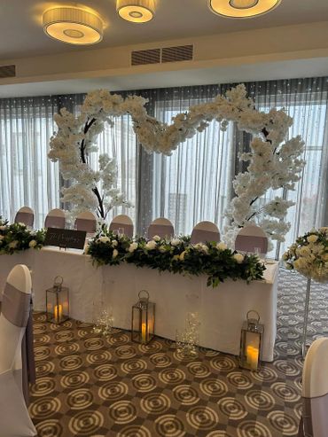 Elegant wedding head table with white floral heart arch and candle lanterns.
