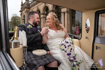 Newlyweds in a vintage car, toasting with champagne and smiling joyfully.