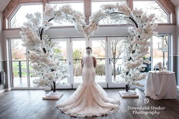 Bride in elegant gown stands under heart-shaped floral arch indoors.