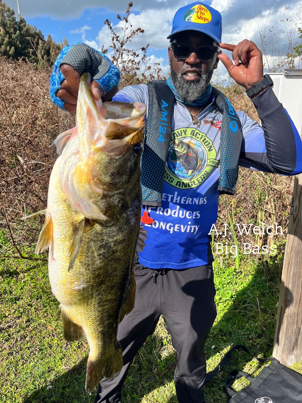 Man proudly holds a large bass fish outdoors in bright sunlight.